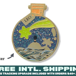 Pode incluir: Pin de esmalte dourado com uma garrafa de vidro com uma rolha de cortiça. A garrafa contém um céu noturno cheio de glitter azul e verde com uma constelação, uma estrela cadente e uma palmeira numa pequena ilha. A garrafa está etiquetada com "LEO".