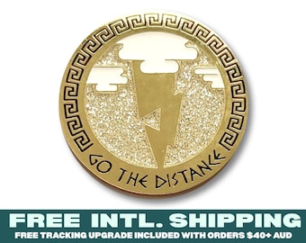 May include: A circular gold-toned metal pin featuring a central lightning bolt design against a glittery background with white clouds. The border displays a Greek key pattern, and the text GO THE DISTANCE is engraved along the bottom edge.