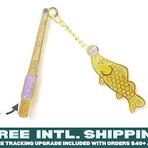 May include: A gold-toned metal charm featuring a fishing rod connected by a small chain to a fish pendant. The rod has sections of purple and orange glitter, while the fish has a yellow body with a honeycomb pattern and a cross symbol.
