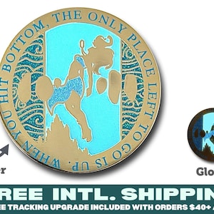 May include: A round gold-toned metal coin featuring a central light blue enamel inlay with a silhouette of a figure floating upward. The border text reads, WHEN YOU HIT BOTTOM, THE ONLY PLACE LEFT TO GO IS UP. The design includes glitter accents and a glow-in-the-dark feature.