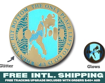 May include: A round gold-toned metal coin featuring a central light blue enamel inlay with a silhouette of a figure floating upward. The border text reads, WHEN YOU HIT BOTTOM, THE ONLY PLACE LEFT TO GO IS UP. The design includes glitter accents and a glow-in-the-dark feature.