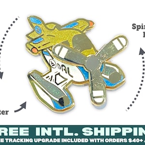 Pode incluir: Um alfinete de avião dourado com uma hélice giratória. O alfinete tem um acabamento brilhante e detalhes em azul e branco. O texto "DAL" é visível no corpo do avião. Um acessório único.