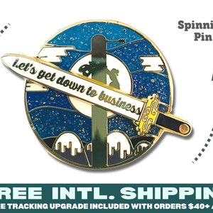 May include: A round, gold-colored spinning pin with a blue and white background, featuring a sword with the text "Let's get down to business." The pin has a detailed design with a night sky and mountain silhouette.