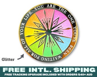 May include: A circular metal coin featuring a rainbow-coloured glitter background with a central black starburst design. The black outer border contains the white text: YOU ARE THE ONE YOU'VE BEEN WAITING FOR ALL OF YOUR LIFE.