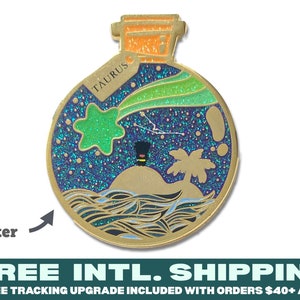 Pode incluir: Pin de esmalte dourado em forma de garrafa com rolha de cortiça. A garrafa contém um líquido brilhante azul e verde com uma estrela, uma constelação e uma palmeira em uma pequena ilha. O texto "TAURUS" está na garrafa.