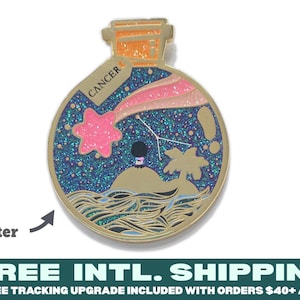 May include: A gold-toned metal enamel pin shaped like a bottle containing a night sky scene with a pink shooting star, a small island with a palm tree, and wavy water. A tag attached to the bottle neck is labeled CANCER. The background features glitter.