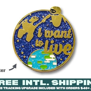 Può includere: Spilla in smalto dorata con sfondo blu glitterato e un'immagine stilizzata di due robot del film Wall-E. Il testo "I want to live" è scritto in lettere dorate.