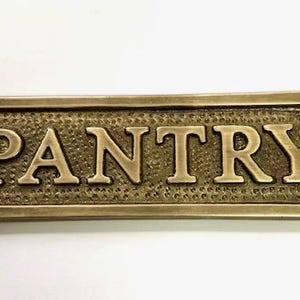 Może przedstawiać: Mosiężny znak z wytłoczonym napisem "PANTRY". Znak ma zaokrąglony prostokątny kształt z ozdobnymi krawędziami i dwoma małymi otworami po obu stronach.