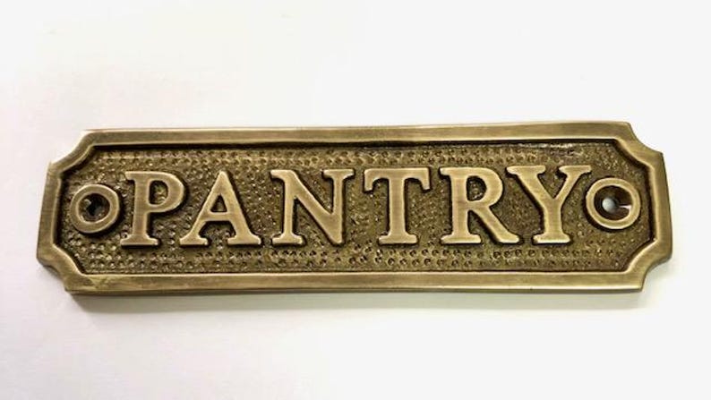 Może przedstawiać: Mosiężny znak z wytłoczonym napisem "PANTRY". Znak ma zaokrąglony prostokątny kształt z ozdobnymi krawędziami i dwoma małymi otworami po obu stronach.