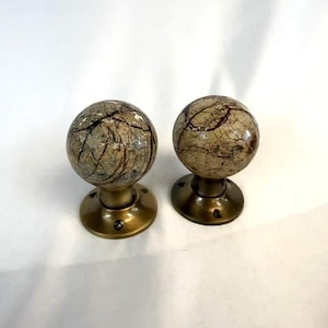 May include: Two spherical door knobs with a mottled beige and brown pattern. They are mounted on a round, brass-coloured base. The knobs have a decorative appearance. The background is plain white.