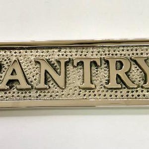 Może przedstawiać: Srebrny metalowy znak z wytłoczonym napisem "PANTRY". Znak ma po dwa otwory po obu stronach do zawieszenia.