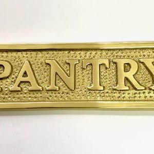 Może przedstawiać: Mosiężny znak z wytłoczonym napisem "PANTRY". Znak ma ozdobną ramkę i dwa małe otwory po obu stronach.