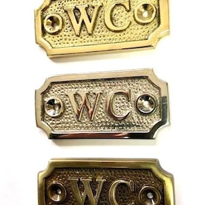 May include: Three decorative WC signs in gold, silver, and bronze tones. Each sign features the letters "WC" in a bold font, with a textured background and decorative edges. The signs have a vintage aesthetic.