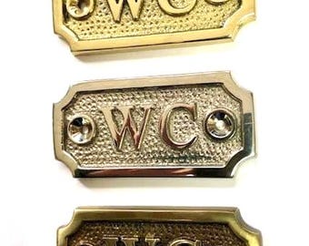 May include: Three decorative WC signs in gold, silver, and bronze tones. Each sign features the letters "WC" in a bold font, with a textured background and decorative edges. The signs have a vintage aesthetic.