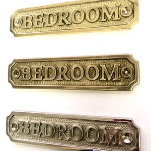 May include: Three brass door signs with the word "BEDROOM" engraved on them. The signs are rectangular with rounded corners and have a textured surface.