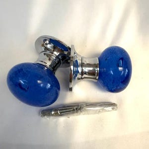 May include: Two door knobs with a bright blue, speckled glass design. The knobs have a chrome base and mounting hardware. The knobs are oval-shaped and are likely for a cabinet or door.