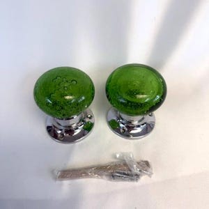May include: Two green glass door knobs with a bubble effect, mounted on shiny silver bases. A small packet of screws is included. The knobs are a vibrant green colour and are likely for a cabinet or door.