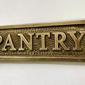 Może przedstawiać: Mosiężny znak z wytłoczonym napisem "PANTRY". Znak ma fakturowaną powierzchnię i zaokrąglone krawędzie.