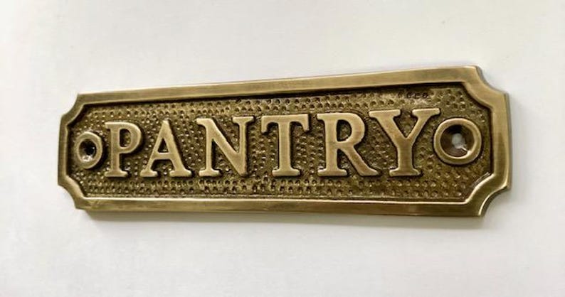 Może przedstawiać: Mosiężny znak z wytłoczonym napisem "PANTRY". Znak ma fakturowaną powierzchnię i zaokrąglone krawędzie.