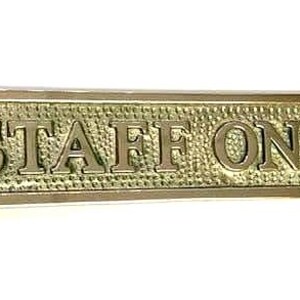 May include: A rectangular brass sign with a textured background and polished edges. The sign features the embossed text "STAFF ONLY" in capital letters. There are two circular screw holes, one on each end of the sign for mounting purposes.