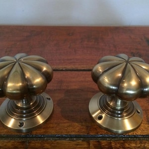 May include: A pair of antique brass door knobs. Each knob features a flower-shaped design with a rounded top and a circular base. The brass has a warm, aged patina. The knobs are mounted on a wooden surface.
