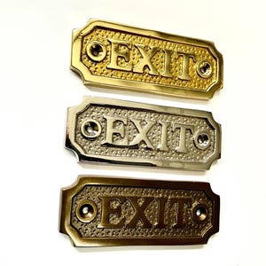 May include: Three metal EXIT signs in gold, silver, and bronze tones. Each sign has the word "EXIT" in raised letters and is mounted on a decorative rectangular plate with rounded edges. The signs are likely for commercial or public spaces.