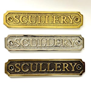 May include: Three rectangular metal signs with the word "SCULLERY" embossed on them. The top sign is gold, the middle is silver, and the bottom is bronze. Each sign has a decorative border and holes for mounting.