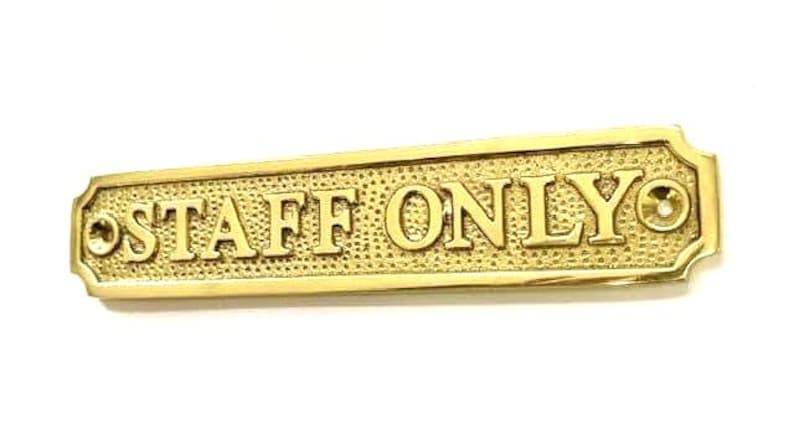 May include: A rectangular brass sign with a textured background and polished edges. The text "STAFF ONLY" is embossed in capital letters across the center. There are two circular screw holes, one on each end of the sign for mounting purposes.