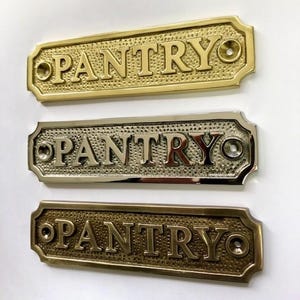 Może przedstawiać: Trzy mosiężne tabliczki na drzwi z wytłoczonym napisem "PANTRY". Tabliczki są w r&oacute;żnych wykończeniach: polerowane mosiądz, satynowy mosiądz i antyczny mosiądz.