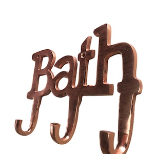 May include: A copper-coloured metal bath sign with three hooks. The sign says "Bath" in a decorative font.