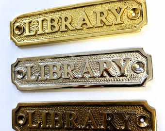 May include: Three rectangular metal door signs with rounded corners, each featuring the word LIBRARY in raised capital letters. The background of each sign has a textured, dimpled finish. The signs are finished in polished brass, chrome, and antique brass, with two screw holes for mounting.