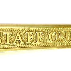 May include: A rectangular brass sign with a textured, hammered background and polished edges. The text "STAFF ONLY" is embossed in a bold, serif font across the center. There are two circular screw holes, one on each end of the sign.