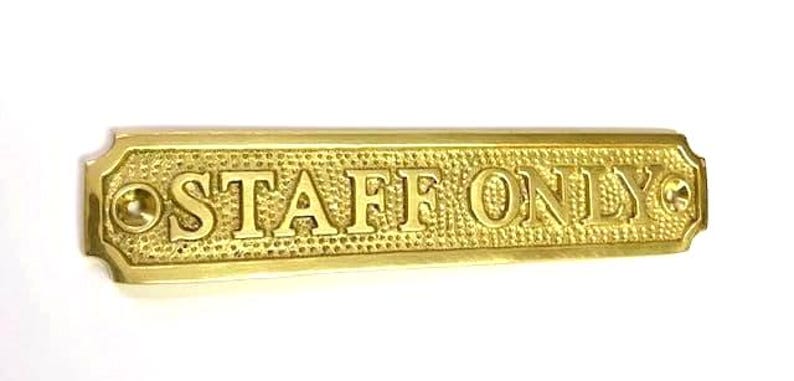 May include: A rectangular brass sign with a textured, hammered background and polished edges. The text "STAFF ONLY" is embossed in a bold, serif font across the center. There are two circular screw holes, one on each end of the sign.
