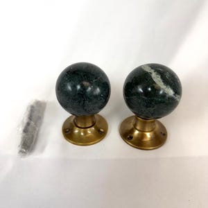 May include: Two dark green marble door knobs with brass bases. The knobs are spherical and have a polished finish. A small packet of screws is visible to the left. Suitable for interior doors.