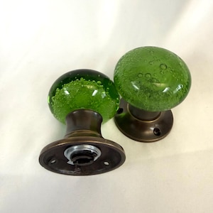 May include: Two green glass doorknobs with a bubble-like texture. The knobs are mounted on bronze-coloured bases with a circular design. The doorknobs are a decorative element for doors.