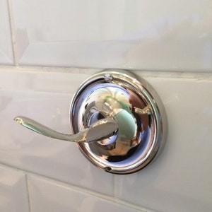 May include: A chrome-plated towel hook mounted on white tiles.