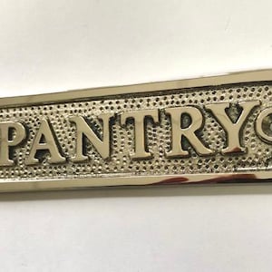 Może przedstawiać: Srebrny, polerowany metalowy znak z wytłoczonym napisem "PANTRY". Znak ma ozdobną ramkę i dwa małe otwory do montażu.