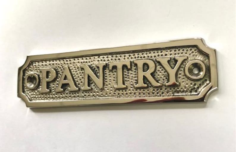 Może przedstawiać: Srebrny, polerowany metalowy znak z wytłoczonym napisem "PANTRY". Znak ma ozdobną ramkę i dwa małe otwory do montażu.