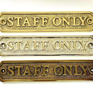 May include: Three rectangular metal signs with rounded corners, each featuring the embossed text "STAFF ONLY" in capital letters. The signs have a textured background and two screw holes on the ends, presented in brass, polished chrome, and antique bronze finishes.