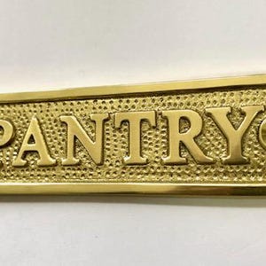 Może przedstawiać: Mosiężny znak z wytłoczonym napisem "PANTRY". Znak ma fakturowaną powierzchnię i dwa małe otwory po obu stronach.
