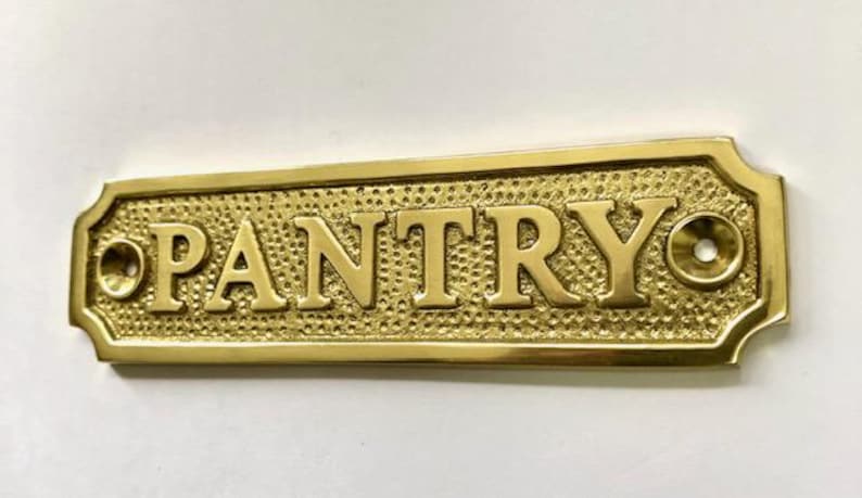 Może przedstawiać: Mosiężny znak z wytłoczonym napisem "PANTRY". Znak ma fakturowaną powierzchnię i dwa małe otwory po obu stronach.