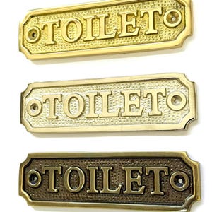 May include: Three rectangular toilet signs in gold, brass, and bronze tones. Each sign has the word "TOILET" in capital letters. The signs have a textured background and are designed with screw holes for mounting.