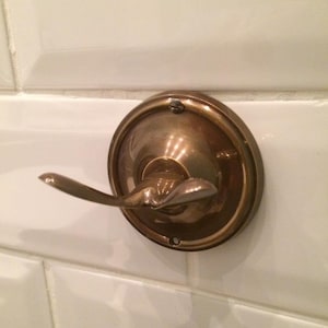 May include: A bronze-coloured wall hook with a circular base and a curved hook extending outwards. The hook is mounted on a white tiled surface, suggesting a bathroom. The hook is made of metal.