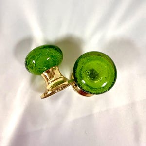 May include: Two green glass door knobs with brass bases. The knobs have a rounded shape with visible air bubbles. The brass bases have a shiny, gold finish. The knobs are likely for a cabinet or door.