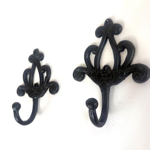 May include: Two black wall hooks with ornate, scrollwork backplates. Each hook features a curved, J-shaped design with a rounded tip. The backplates have symmetrical, open-work patterns with swirling details, presented in two different sizes.