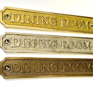 May include: Three rectangular "DINING ROOM" signs in gold, silver, and bronze tones. Each sign has a textured surface and decorative edges with screw holes. The signs are for a dining room.