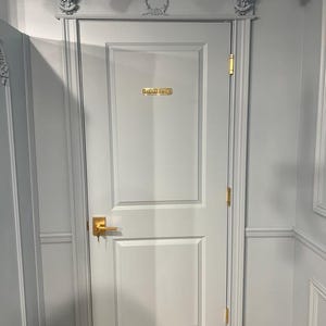 May include: A light gray door with two rectangular panels, featuring a gold-colored handle, gold hinges, and a gold plaque that reads "STAFF ONLY". The door frame includes decorative molding with carved floral details and a central wreath motif.