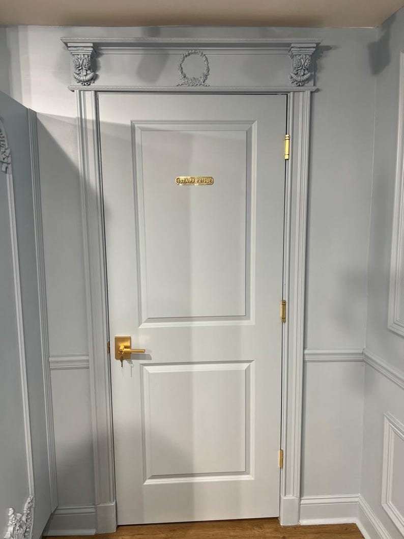 May include: A light gray door with two rectangular panels, featuring a gold-colored handle, gold hinges, and a gold plaque that reads "STAFF ONLY". The door frame includes decorative molding with carved floral details and a central wreath motif.