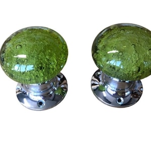 May include: A pair of green glass door knobs. Each knob has a textured, bubble-like appearance and sits on a polished, chrome-effect base. The glass is translucent. The knobs are suitable for doors.