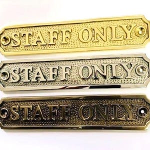 May include: Three rectangular metal signs with rounded corners, each featuring the embossed text "STAFF ONLY" in capital letters. The signs have a textured background and a screw hole on each end, presented in polished brass, chrome, and antique bronze finishes.